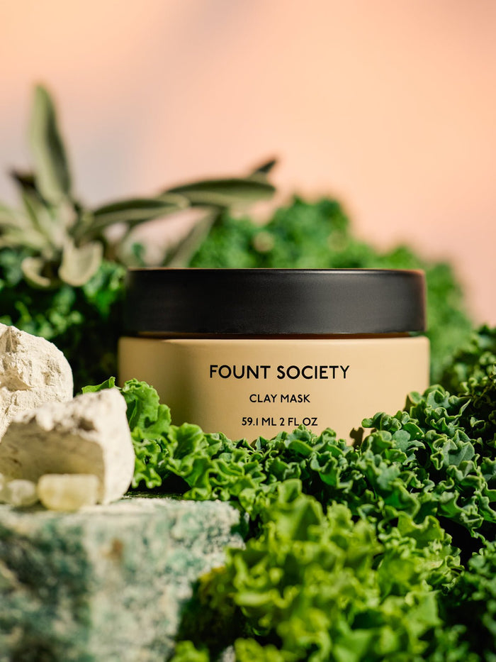 A Cozy Earth Clay Mask container is nestled among lush greenery and rocks, with a blurred pastel background. |Size:Full Size