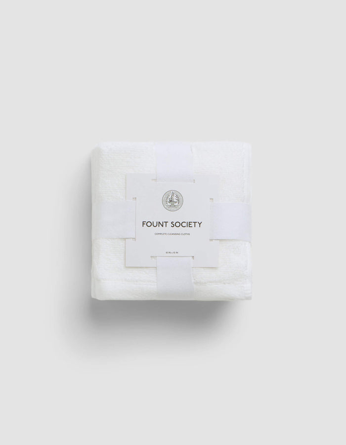 A neatly folded white Complete Cleansing Cloth by Cozy Earth, wrapped with a white paper band featuring the brand name and a small circular logo, set against a plain light gray background.|Color:White