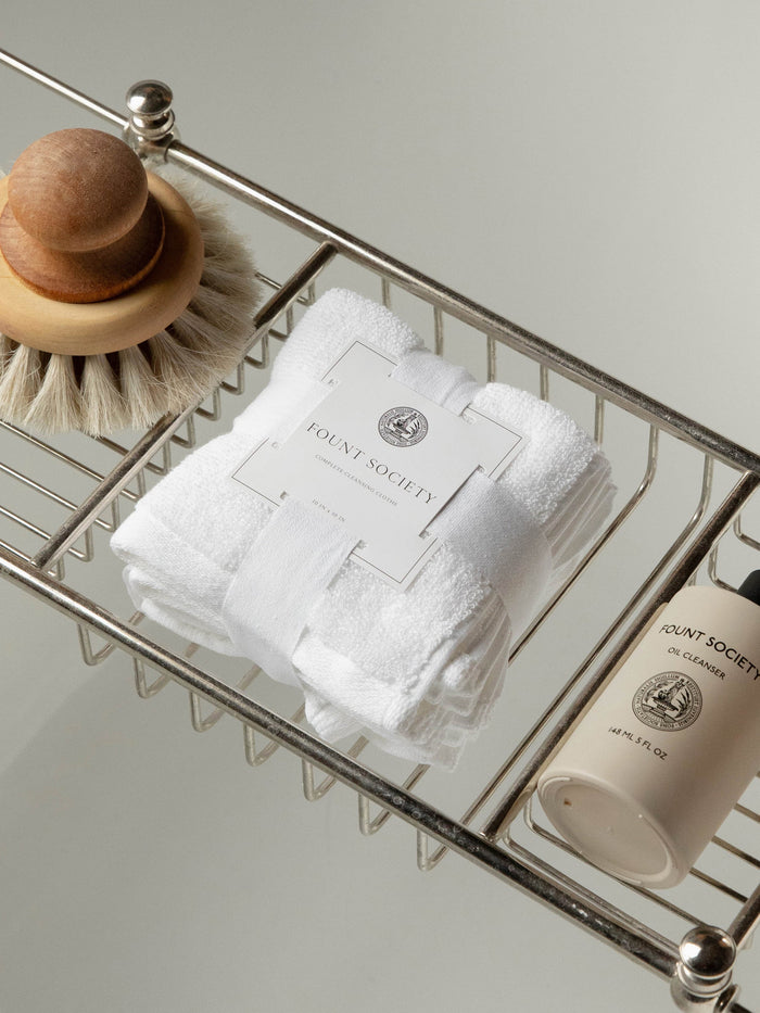 A wire rack holds toiletries: a wooden body brush with bristles on one side, a folded white towel with a "Cozy Earth" label, and a cylindrical container labeled "Complete Cleansing Cloths." The wire rack appears to be part of a shower or bath setup.|Color:White