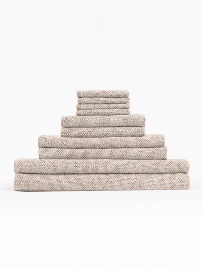 The Complete Nantucket Bath Bundle by Cozy Earth, featuring beige textured towels in various sizes, is neatly stacked pyramid-style on a white background.|Color:Heathered Sand
