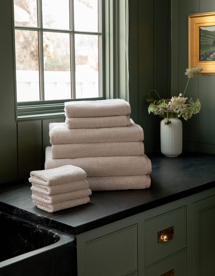 The Complete Nantucket Bath Bundle by Cozy Earth, in beige, is neatly stacked on a black countertop near a window with a vase of flowers and pale green cabinetry in the background.|Color:Heathered Sand