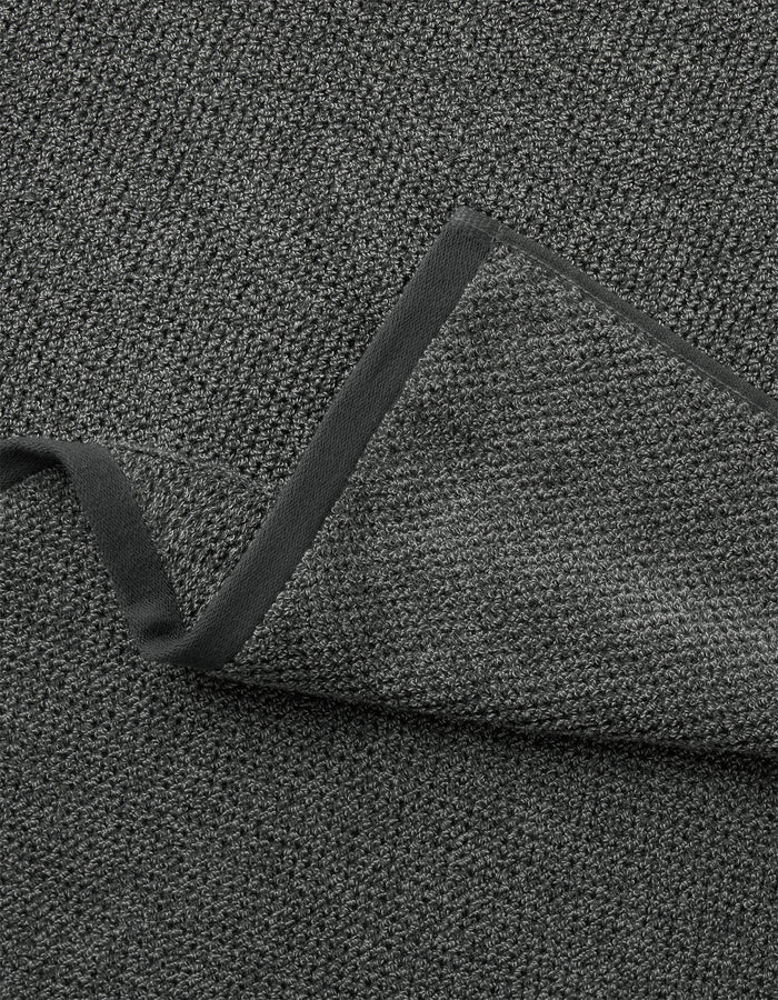 A close-up of the Cozy Earth Nantucket Bath Towel Set in dark gray, showing its textured fabric and black trim, with one corner folded to reveal its thickness.|Color:Graphite