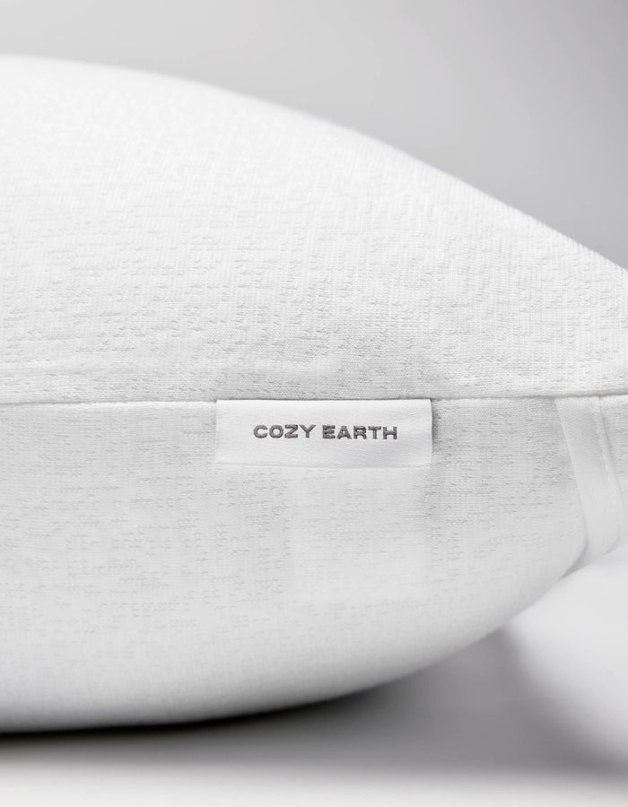 A close-up of the Cozy Earth Cooling Pillow Protector showcases its textured white fabric and a small tag reading "COZY EARTH" in gray. The all-white background creates a clean, minimalist aesthetic.