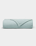 The Cozy Earth Cotton Gauze Blanket is neatly folded in light blue with a textured, grid-like pattern on a plain white background. One corner is turned down to reveal the inner side.|Color:Tide