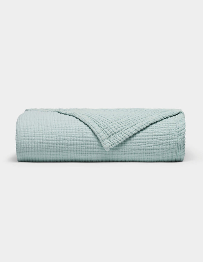 The Cozy Earth Cotton Gauze Blanket is neatly folded in light blue with a textured, grid-like pattern on a plain white background. One corner is turned down to reveal the inner side.|Color:Tide
