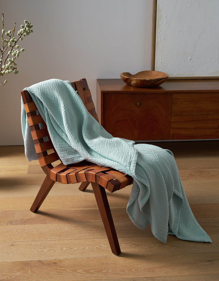 A wooden woven chair draped with the Cozy Earth Cotton Gauze Blanket in light blue sits on a wooden floor beside a sideboard topped with a decorative bowl and framed abstract art.|Color:Tide