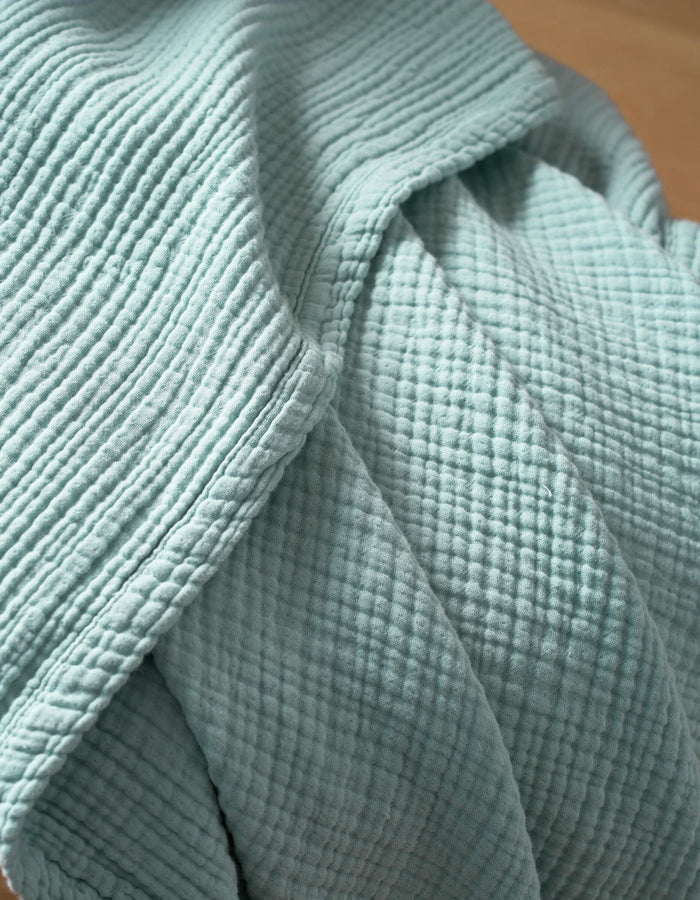 A close-up of Cozy Earth's Cotton Gauze Blanket highlights its textured, light blue woven fabric with a grid pattern, soft folds, and stitched seams.|Color:Tide