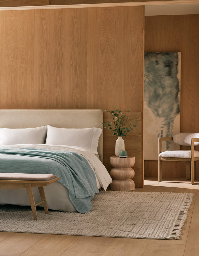 A modern bedroom with light wood-paneled walls, a cream bed topped with Cozy Earth's Cotton Gauze Blanket, white and light blue bedding, a layered side table with greenery, a beige bench, and a soft woven rug.|Color:Tide