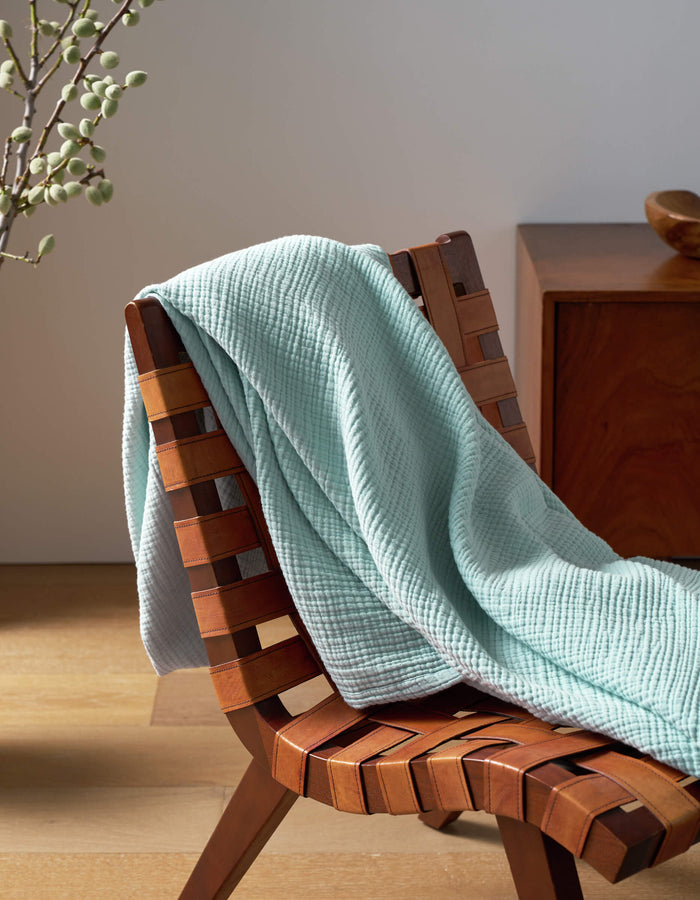 A Cozy Earth Cotton Gauze Blanket in light blue is draped over a woven wooden chair, next to a wooden side table with a vase of branches, all set on a light wood floor.|Color:Tide