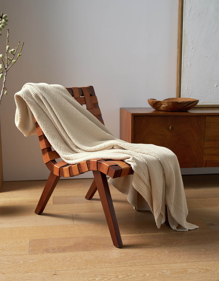 A Cozy Earth Cotton Gauze Blanket in beige is draped over a wooden woven chair beside a modern sideboard, accented by a decorative bowl and abstract wall art in a sunlit, minimalist room.|Color:Coconut