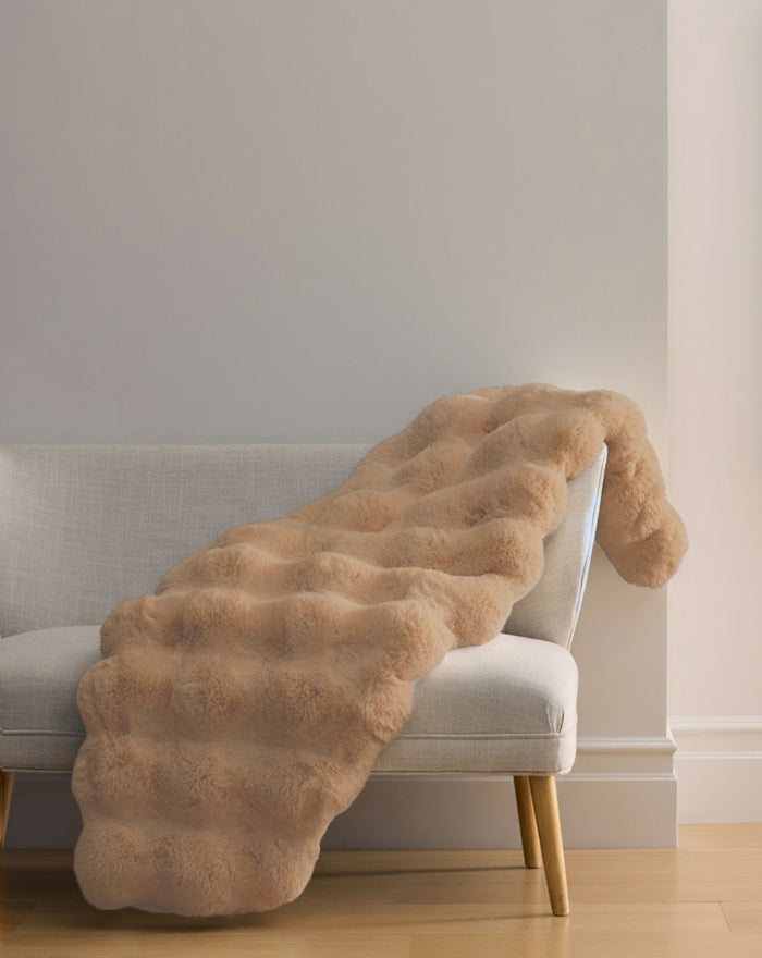 The Cozy Earth Double Bubble Cuddle Blanket, a light beige faux fur throw with a wavy texture, is draped over the arm of a modern light gray sofa in a minimalist room with light wood floors and neutral walls.|Color:Brandy