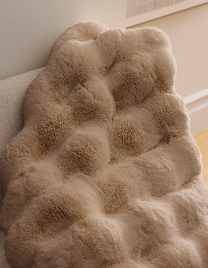 A close-up of the Double Bubble Cuddle Blanket by Cozy Earth, featuring soft ivory beige textured faux fur, draped over a light couch and illuminated by natural light for a cozy look.|Color:Brandy