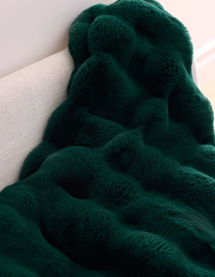 The Double Bubble Cuddle Blanket by Cozy Earth, in dark green plush faux fur with a wavy texture, is draped over the back of a light-colored sofa.|Color:Adrift