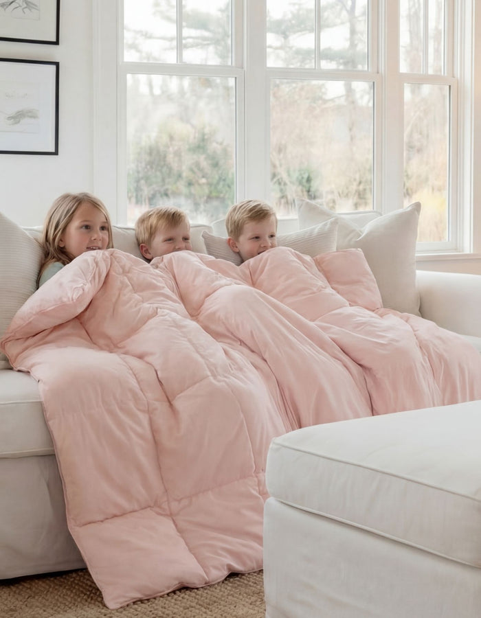 Three young children sit on a white couch, wrapped together under the Cozy Earth Down Alternative Throw in light pink. They look cozy as they relax in front of large windows bathed in natural light. |Color:Petal