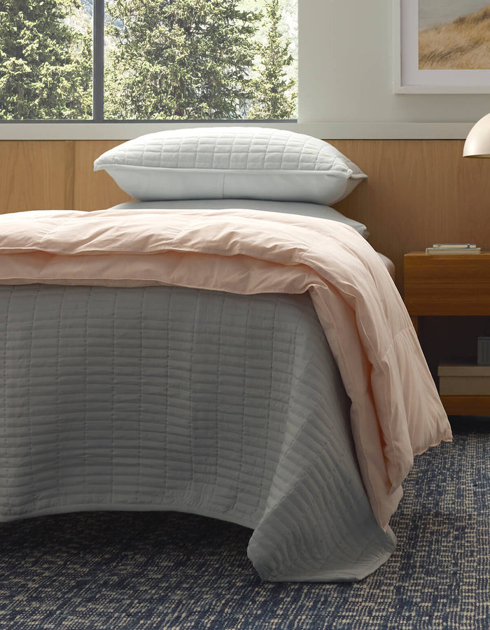 A bed features the Cozy Earth Down Alternative Throw in light gray, a pale pink comforter, and two stacked pillows. Nearby is a wooden nightstand, a blue patterned carpet, and a window with views of green trees.|Color:Petal