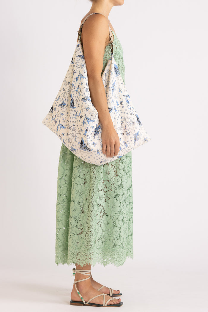 Woman wearing an apple floral lace maxi dress with a cream tote bag with features a blue limited edition percussion print