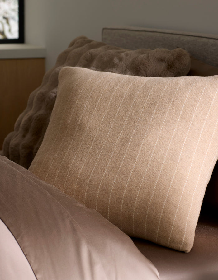 A close-up of a neatly made bed features Cozy Earth's Florence Pillow, a beige striped accent placed in front of a textured brown cushion. Soft brown bedding and a window with blurred outdoor light complete the cozy scene. |Color:Brandy