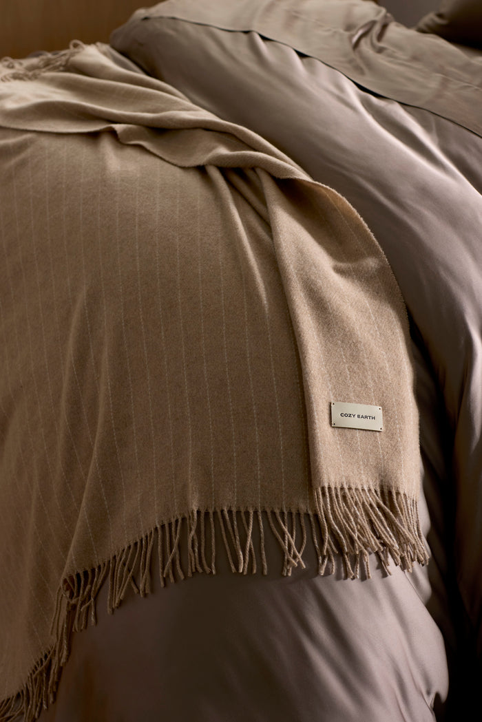 The Florence Throw by Cozy Earth, a soft beige blanket with fringed edges and a "COZY EARTH" label, is draped over taupe bedding on a neatly made bed.|Color:Brandy