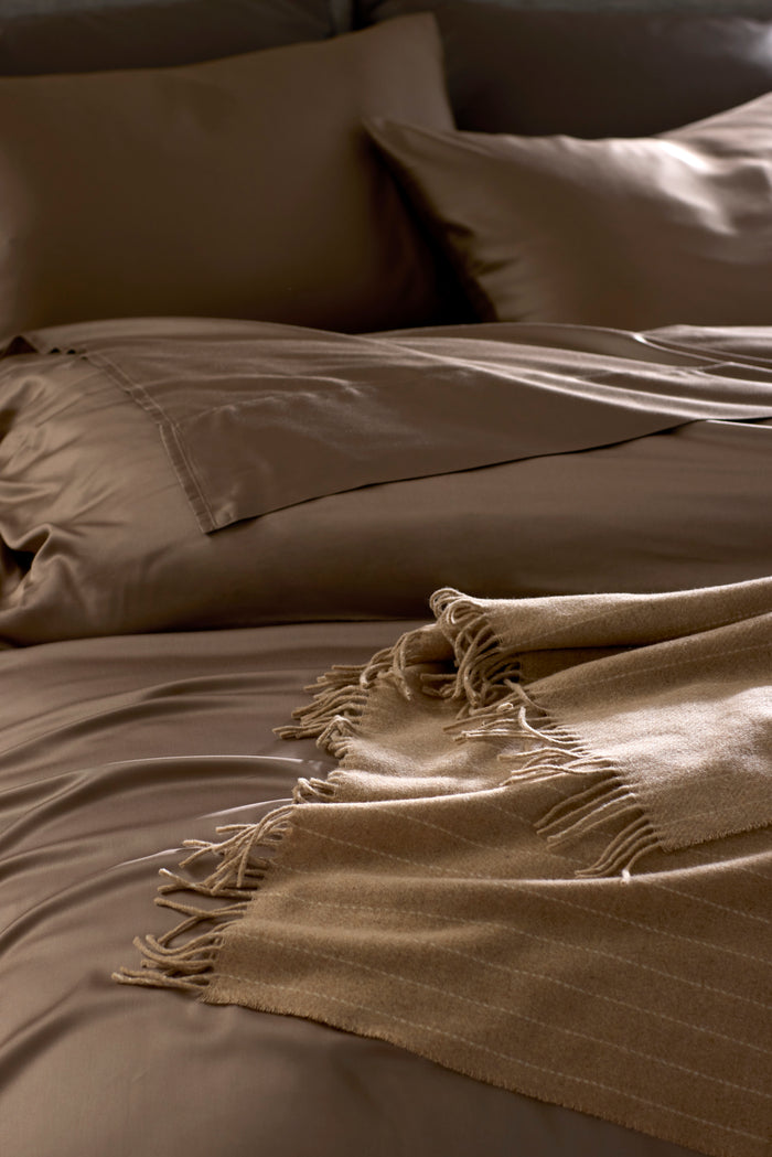 A neatly made bed with brown sheets, matching pillowcases, and the Cozy Earth Florence Throw in beige, featuring a fringed edge and folded at the corner. The bedding creates a smooth, inviting, and warm atmosphere.|Color:Brandy