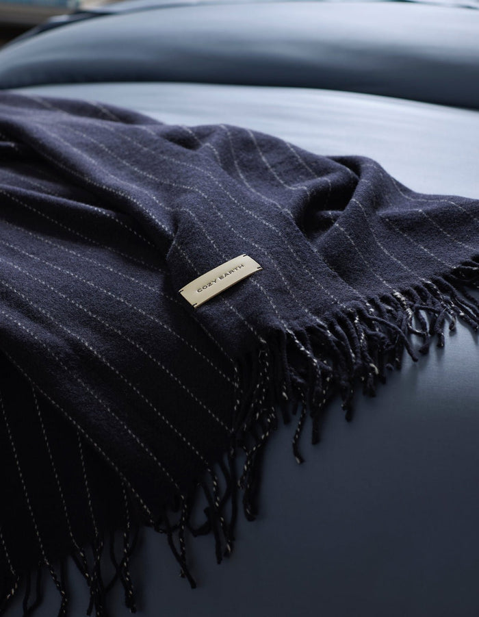 A dark blue, pinstriped Florence Throw by Cozy Earth with fringe is draped over a smooth, light blue surface. A rectangular label showing "COZY EARTH" is attached to the blanket. |Color:Navy