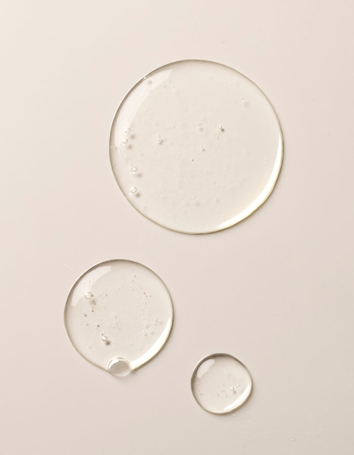 Three clear drops of Gel Cleanser by Cozy Earth on a beige surface. The drops vary in size, ranging from a large drop at the top to smaller ones below, showcasing a glossy and slightly reflective texture.|Size:Full Size