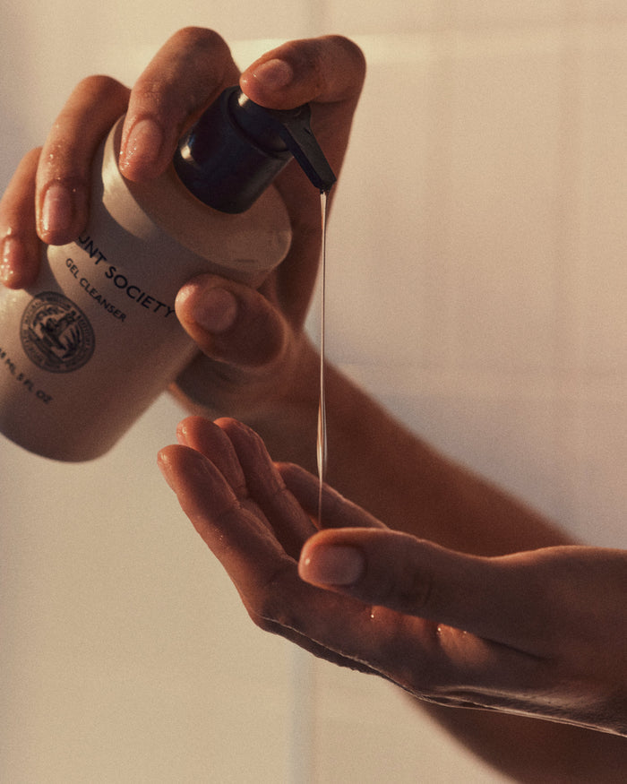 Close-up of hands dispensing Cozy Earth's Gel Cleanser from a pump bottle. One hand is holding the bottle and pressing the pump, while the other hand is positioned below to catch the gel. The background is softly lit with warm tones.|Size:Full Size