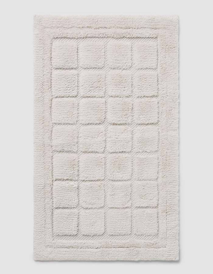The Cozy Earth Grid Bath Mat is a rectangular, off-white mat with a raised grid design and a thick, fluffy border.|Color:Light Grey