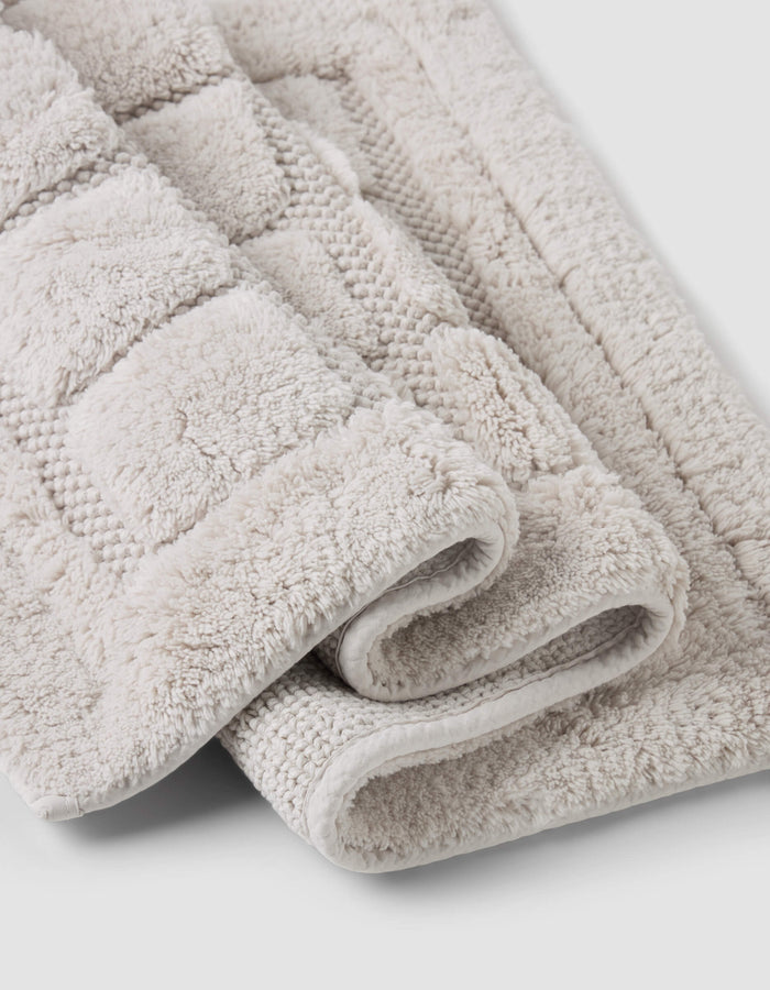 A close-up of two folded Grid Bath Mats by Cozy Earth, featuring a soft, plush surface and geometric texture in beige, displayed on a light gray background.|Color:Light Grey