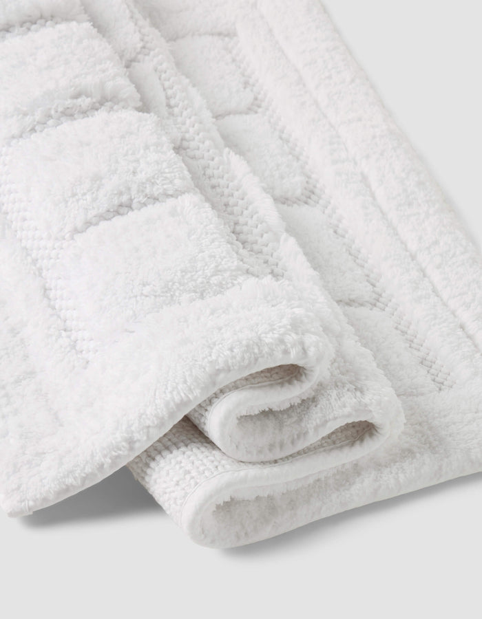 A close-up of the Folded Grid Bath Mat by Cozy Earth, featuring a white textured geometric pattern, displayed on a light gray background.|Color:White