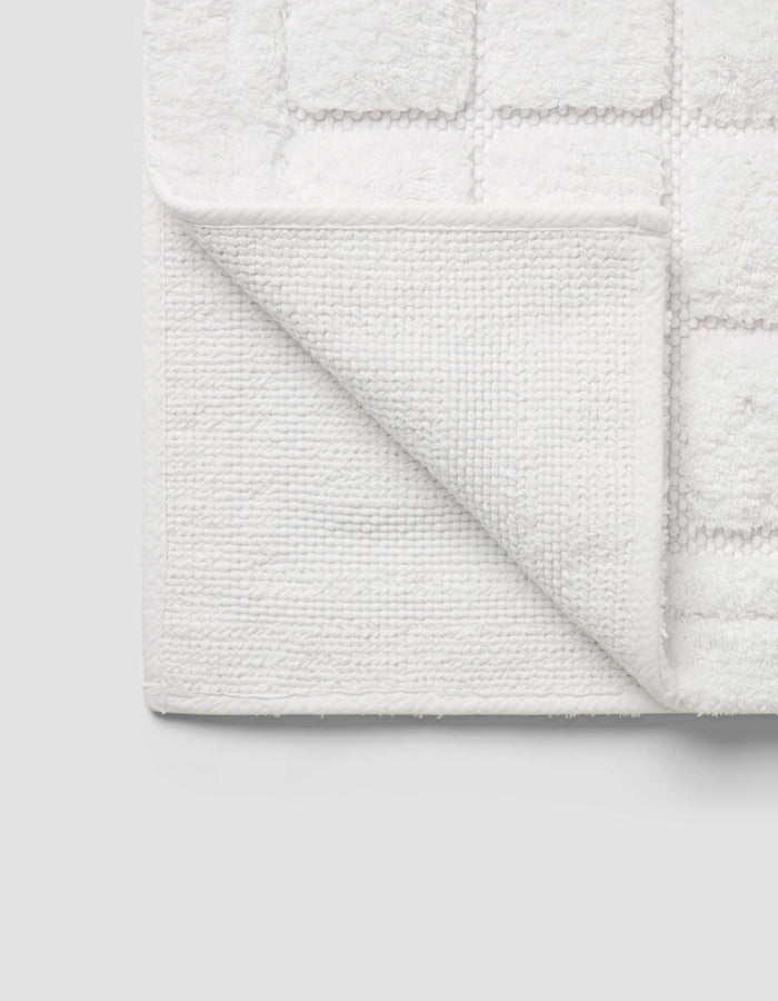 The Cozy Earth Grid Bath Mat, featuring a textured square pattern and woven underside, is displayed with one corner folded on a light gray background.|Color:White