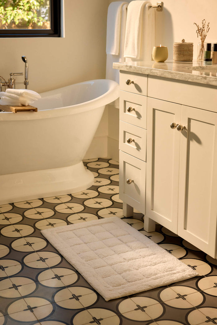 A bathroom with a patterned tile floor, a white freestanding bathtub, a white vanity with drawers, Cozy Earth's Grid Bath Mat, and neatly folded towels on the counter and towel bar. Sunlight filters in from a window.|Color:White