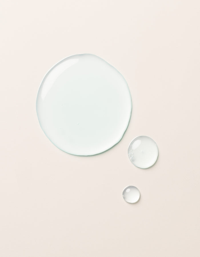 A large clear droplet and two smaller droplets of Cozy Earth Hydrating Serum rest on a smooth, light beige surface. The glossy, semi-transparent droplets reflect light slightly on their surfaces.