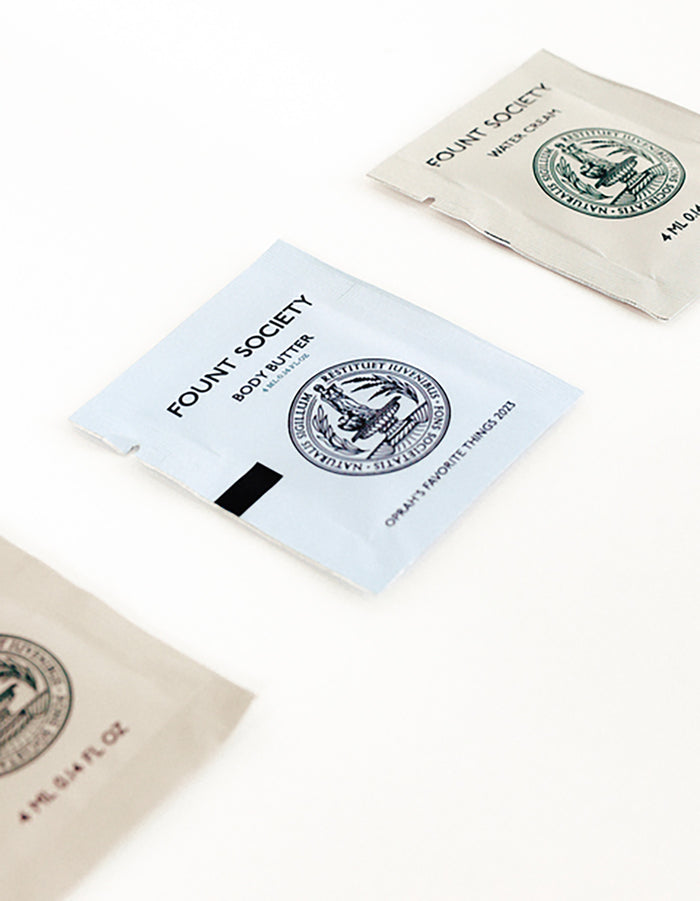 Three small, flat packets from the Cozy Earth Fount Society Intro Kit are arranged on a white surface. The nearest packet reads "BODY BUTTER" and each displays a circular emblem with product details in minimal black text.