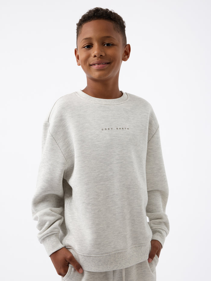 A smiling boy wearing the Kid's CityScape Crewneck by Cozy Earth, featuring "COZY EARTH" printed on it, stands against a plain white background. |Color:Heather Grey