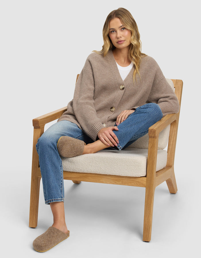 A woman with long blonde hair sits relaxed on a wooden armchair, wearing blue jeans and Cozy Earth’s Lakehouse Clog slippers. One leg is crossed over the other, and she looks calmly at the camera.|Color:Chestnut