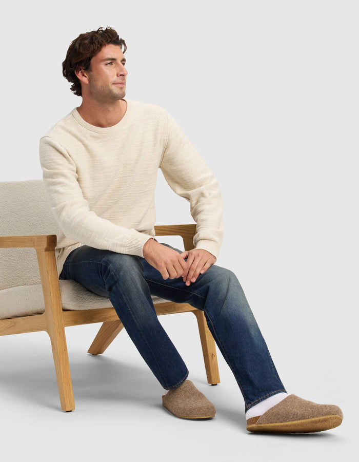 A man in a cream sweater and blue jeans sits on a light wooden chair, wearing Cozy Earth Lakehouse Clogs and gazing to the side with a neutral expression.|Color:Chestnut