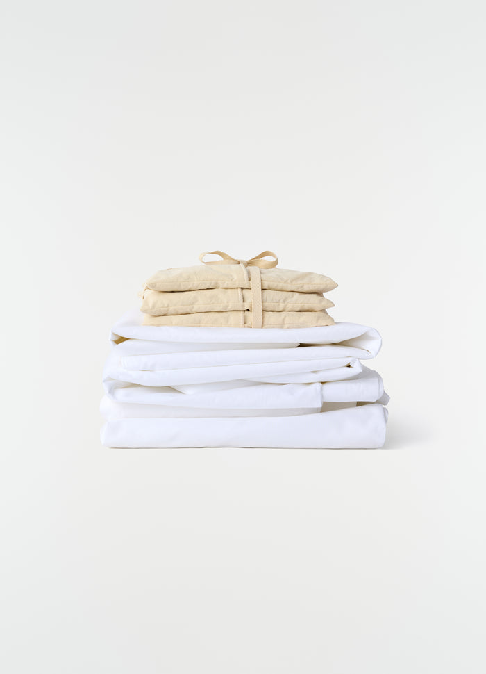 Lavender cushion in beige cotton tied with strap stacked on white linen sheets LEMAIRE plain white background