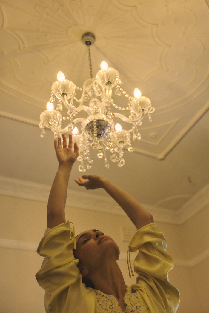 Woman wearing a vanilla lace-lined long sleeve top reaching up towards a chandelier