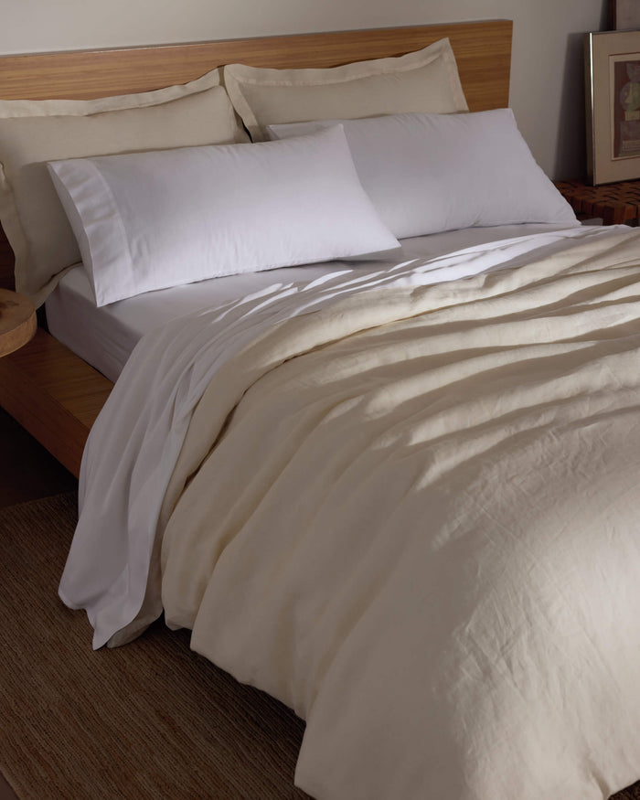 A bed features the Cozy Earth Linen Duvet Cover, white pillows, and a wooden headboard. A bedside table adds warmth to the softly lit room, creating a cozy and inviting atmosphere.|Color:Flax