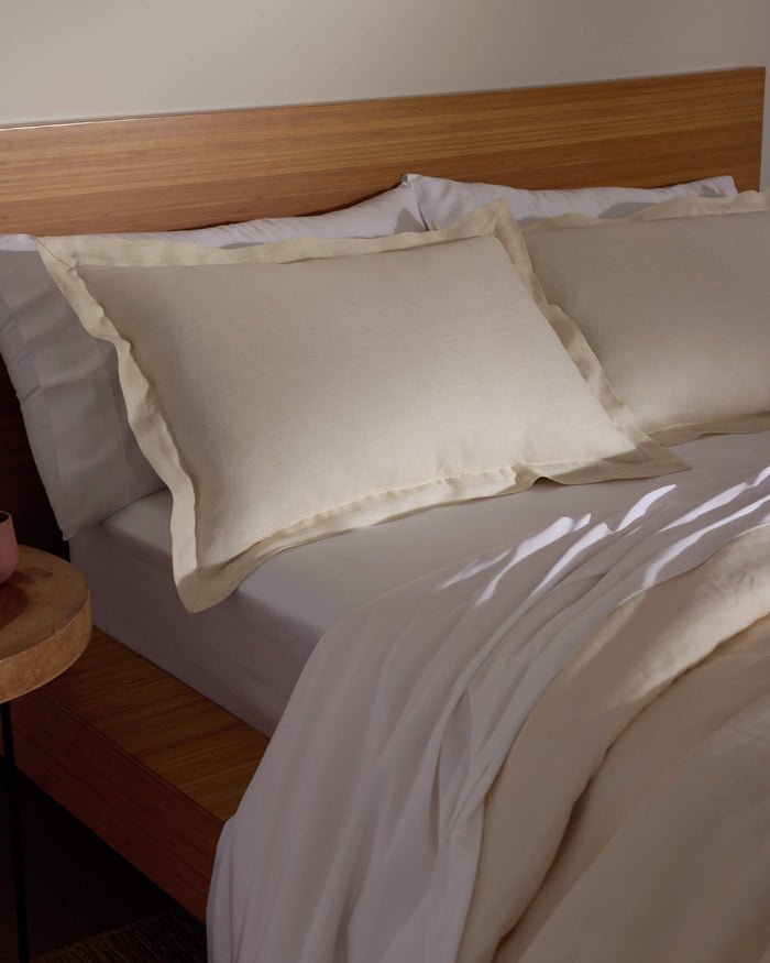 A bed featuring Cozy Earth's Linen Shams in cream, paired with a matching comforter, sits against a wooden headboard and nightstand, all bathed in soft natural light.|Color:Flax