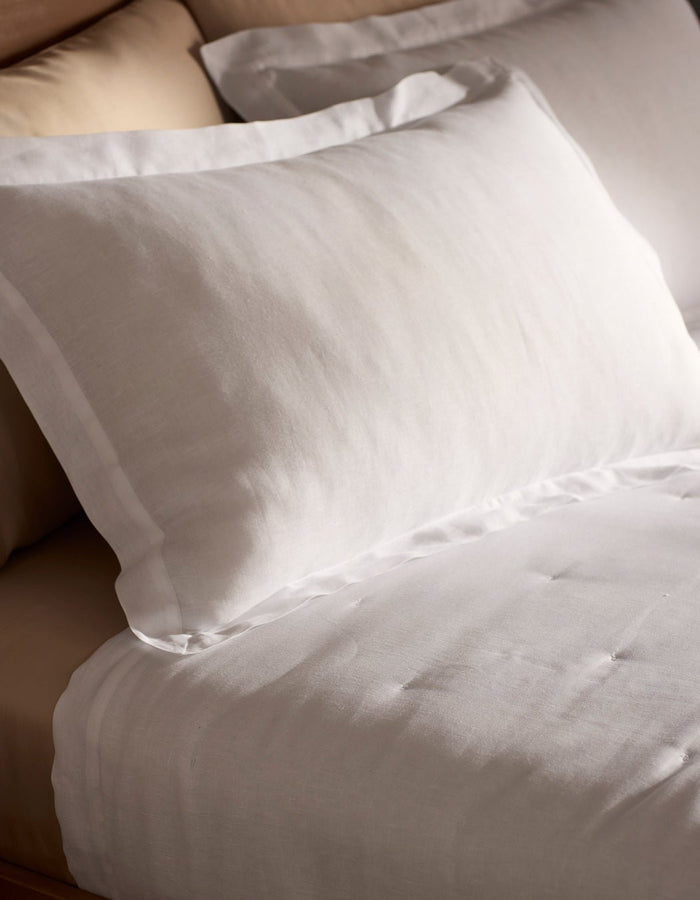 Close-up of a neatly made bed with Cozy Earth Linen Shams in white, showcasing their smooth, crisp texture. The bed features a beige headboard and looks soft and inviting.|Color:White
