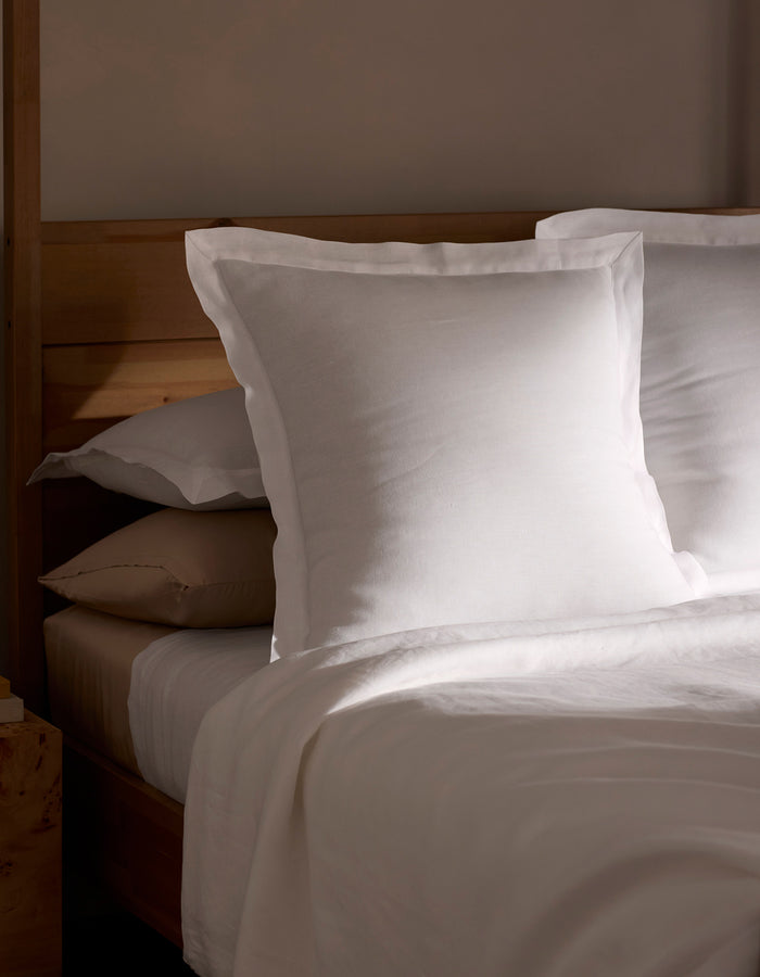 A neatly made bed is shown in soft natural light, featuring Cozy Earth Linen Shams stacked behind crisp white pillows and sheets, all set against a wooden headboard.|Color:White