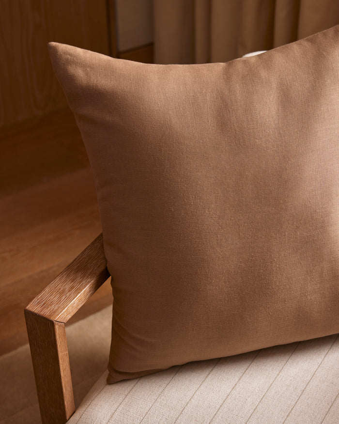 A Cozy Earth Linen Throw Pillow sits on a light beige upholstered chair with a wooden armrest, in a softly lit room featuring wooden flooring and neutral curtains.|Color:Maple|Size:24" x 24"