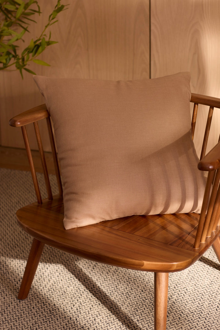 A Cozy Earth Linen Throw Pillow rests on a wooden chair with a spindle backrest, bathed in soft sunlight. A textured rug and leafy plant add coziness to the minimalist space.|Color:Maple|Size:20" x 20"