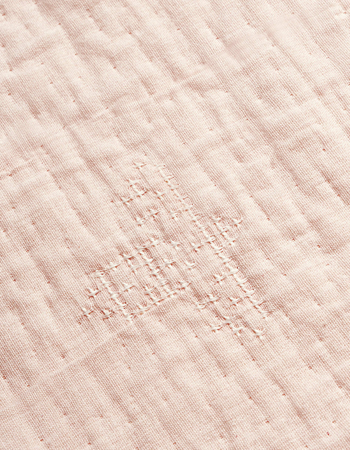 A close-up of the Cozy Earth Little Bee Matelasse Baby Blanket shows its white fabric with a grid-like, hand-stitched repair in matching thread, highlighting the textured and slightly uneven surface.|Color:Petal