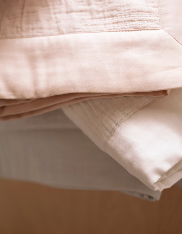 A close-up of the Cozy Earth Little Bee Matelasse Baby Blanket, featuring neatly folded layers of soft beige and white fabric that highlight its cozy texture and detailed edges.|Color:Petal