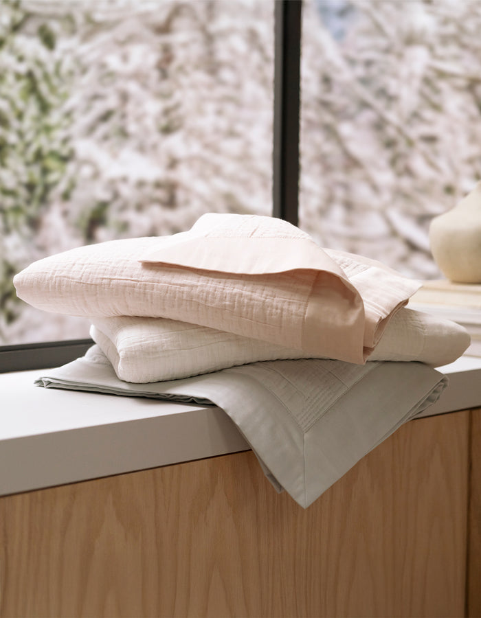 Two Cozy Earth Little Bee Matelasse Baby Blankets in soft pastel hues are folded and stacked on a wooden cabinet by a window, with blurred greenery visible outside.|Color:Vapor