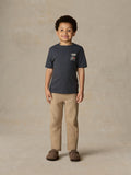 Child wearing a blue graphic shirt and beige pants against a beige background