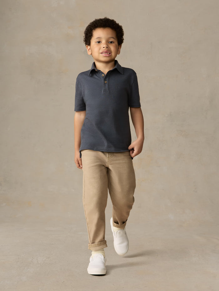Child wearing a blue polo shirt and beige pants against a beige background