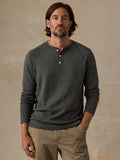 A man wearing a dark green long-sleeve henley shirt and khaki pants, standing against a textured beige studio background.